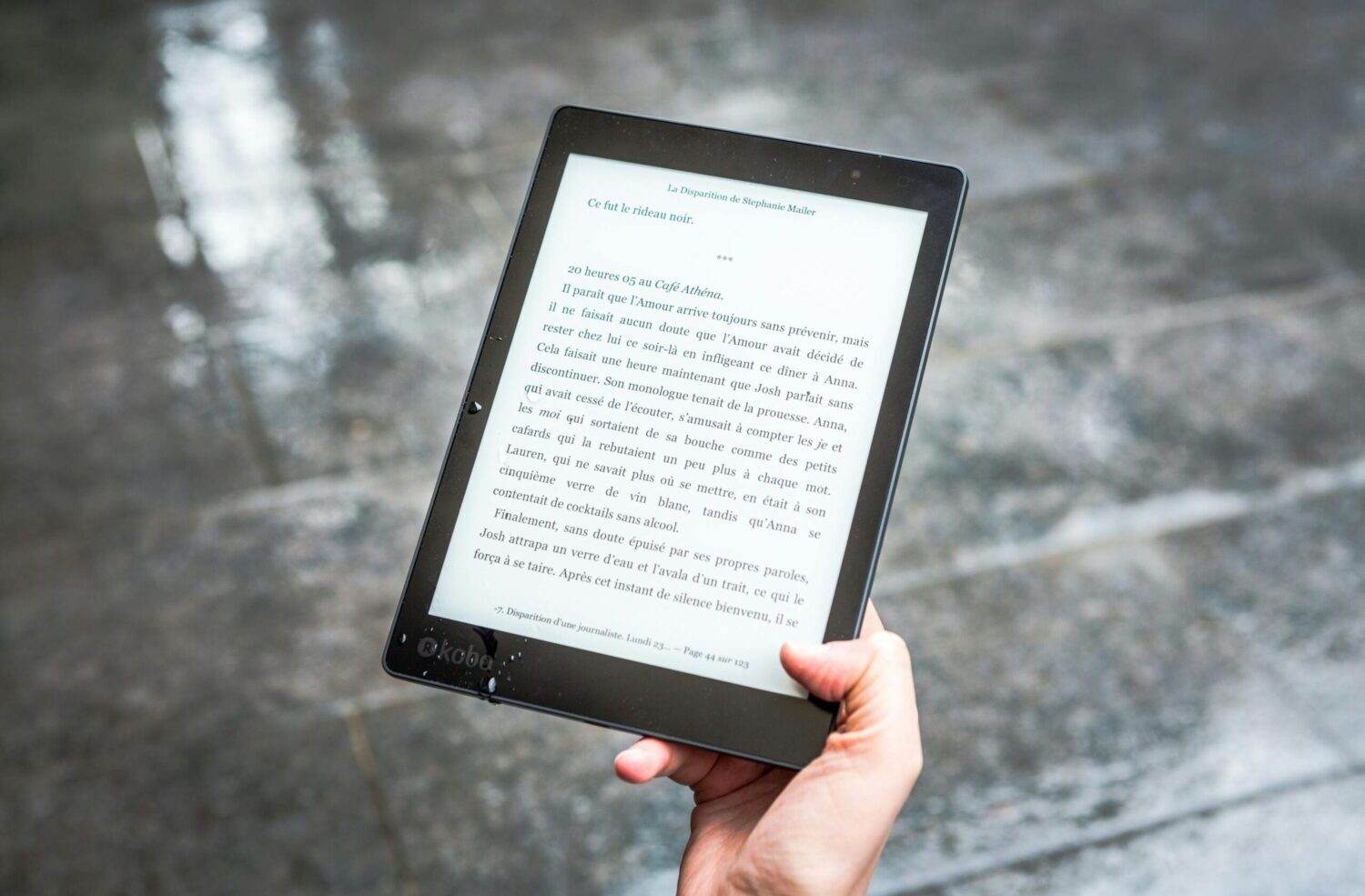 A person holds an e-reader displaying text against a wet pavement background.